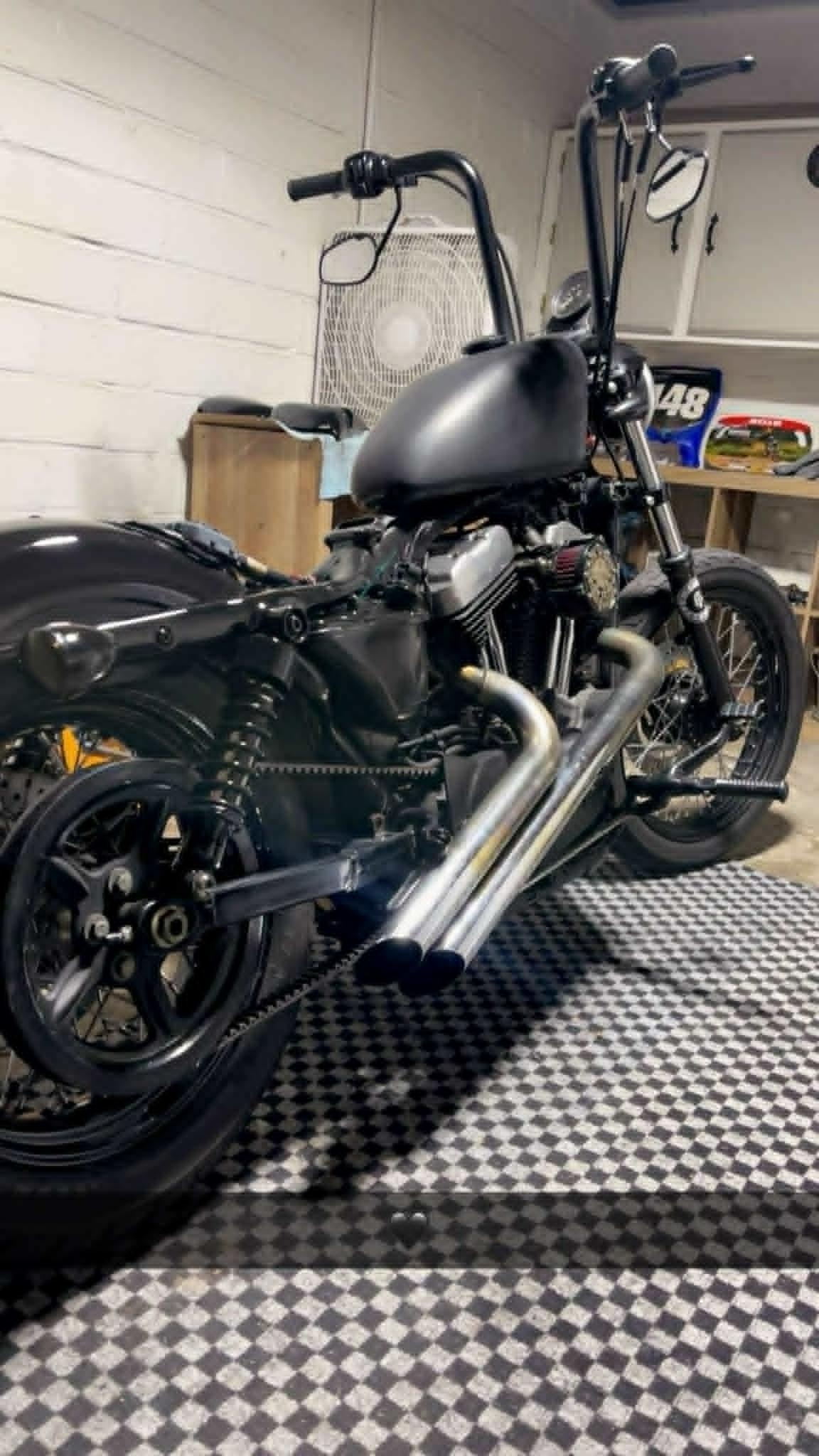Black Sportster bobber inside the shop — mini ape bars, twin chrome pipes, white walls