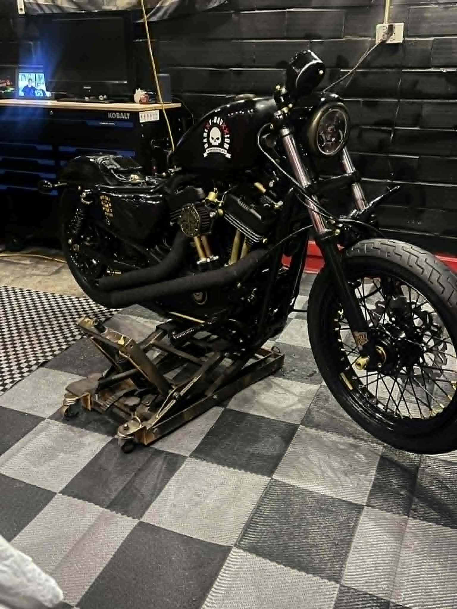 Black Sportster bobber on lift in the shop — Davxxion skull tank, gold accents, checkered floor